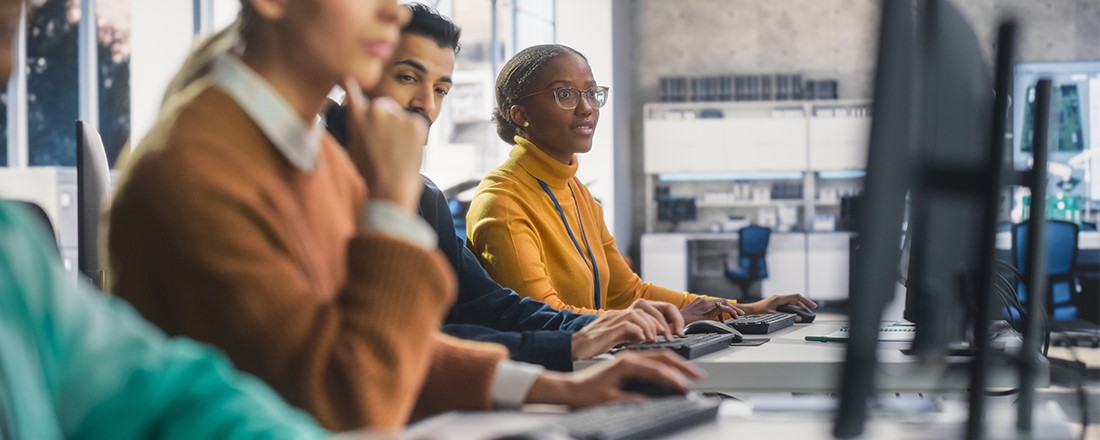 Vier Personen sitzen nebeneinader vor Computern und arbeiten Konzentriert. Die Person im Fokus lächelt.